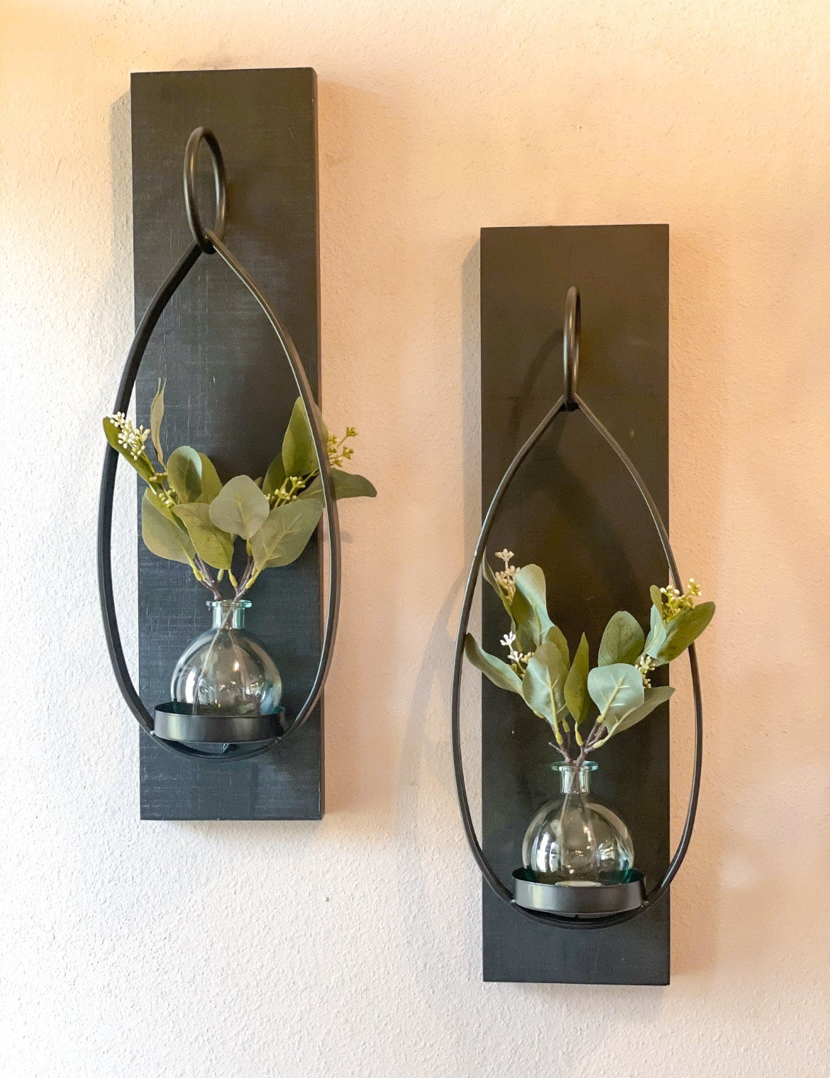 Two black wooden sconces with a metal tear-drop shaped candle holder. There is a round blue glass vase on each candle holder with eucalyptus in each vase making 2 complete sconces.