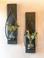 Two black wooden sconces with a metal tear-drop shaped candle holder. There is a round blue glass vase on each candle holder with eucalyptus in each vase making 2 complete sconces.